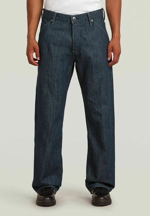 Mann steht, trägt locker sitzende dunkelblaue Jeans und schwarze Schuhe auf hellem einfarbigem Hintergrund.