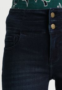 Jeans de mezclilla oscura con una cintura alta, dos botones dorados en la cinturilla y un bolsillo lateral. La tela tiene una textura suave y ligeramente elástica.