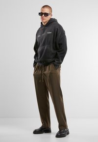 Sweat à capuche en polaire noire avec poche frontale et détail graphique, associé à un pantalon de jogging vert olive et des chaussures noires imposantes. Accessoires : lunettes de soleil noires.