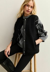 Svart, fluffig väst med svart, vadderad insida, bärs över en grå stickad tröja med diamantmönster. Modell sitter på en pall.