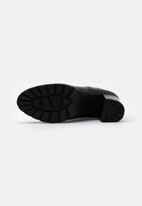 Zapato de cuero negro con una suela de goma texturizada que presenta surcos profundos, puntera redondeada y discretos acentos metálicos a lo largo de la parte superior.