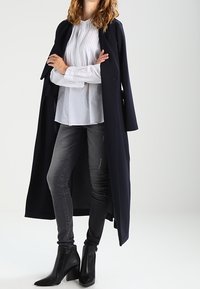 Long manteau bleu marine porté sur un chemisier blanc texturé, assorti à un jean skinny gris et des bottines noires à bout pointu.