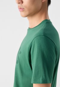 Vihreä puuvillapaita, jossa on pyöreä kaula-aukko, lyhyet hihat ja sileä kangas. Edessä on hillitty printti tai yksityiskohta.