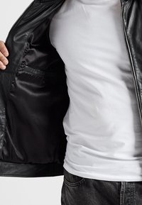 Černá kožená bunda s hladkým povrchem, zipovým zapínáním a vnitřní kapsou, nošená přes jednoduché bílé tričko a tmavé džínové kalhoty.