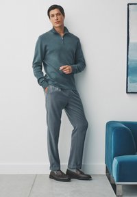 Pull zippé en bleu sarcelle de poids moyen associé à un pantalon texturé gris et des chaussures marron. Design minimaliste sur fond clair.