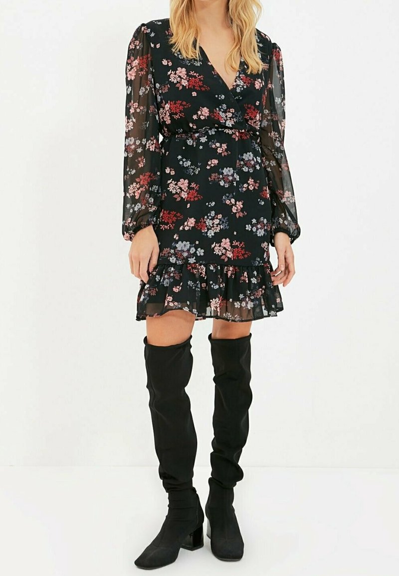 Robe noire à motifs floraux avec des manches longues transparentes et un ourlet volanté. Elle présente un décolleté en V profond et une taille cintrée, ornée de motifs floraux colorés.