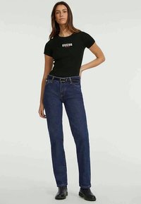 Camiseta justa preta com o logótipo branco "GUESS", combinada com jeans de perna reta azul-escura. Modelo usa botas de tornozelo pretas.