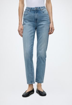 Ben iført lyseblå højttaljede, lige jeans kombineret med sorte flade sko og en hvid top mod en ensfarvet baggrund.