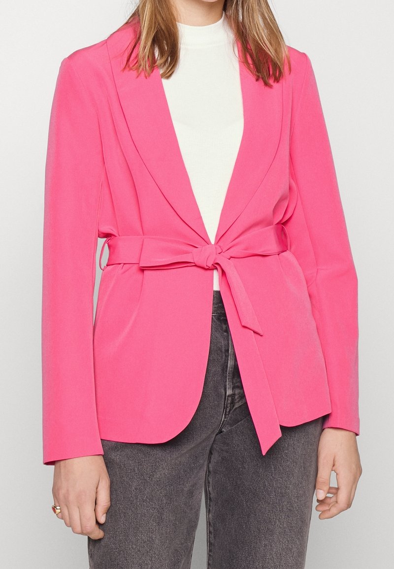 Blazer rose vif à manches longues, au design ajusté, avec un détail de nouage à l'avant. Porté par-dessus un haut blanc et associé à un pantalon gris foncé.