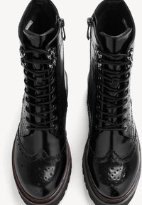 Bottes à lacets brogue en cuir noir brillant avec semelles épaisses bordées de brun, fermetures éclair latérales et perforations détaillées sur les bouts. 