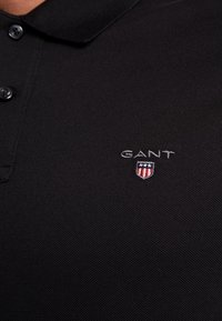 Polo noir avec un col boutonné présentant un petit logo GANT et un emblème en forme de bouclier brodé sur la poitrine gauche.