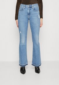 Jean évasé bleu taille haute, délavé clair, avec des détails usés et des ourlets effilochés, assorti à des chaussures noires à talons.