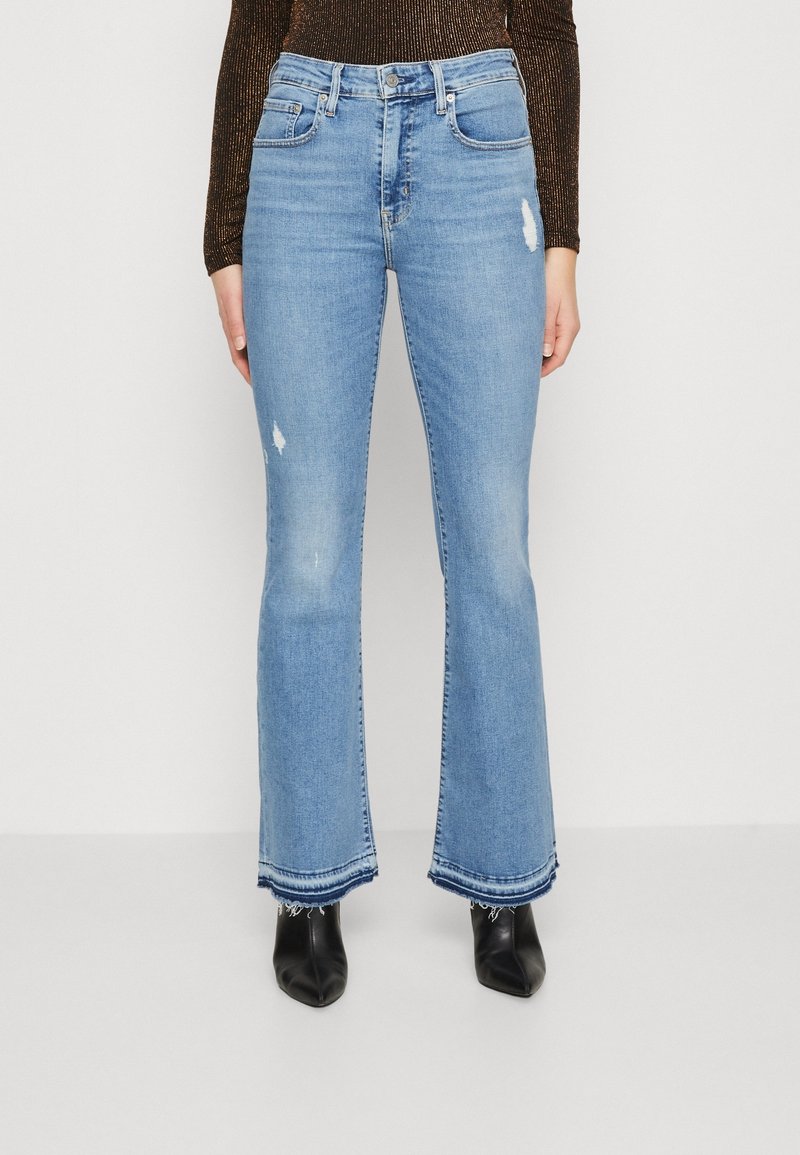 Jean évasé bleu taille haute, délavé clair, avec des détails usés et des ourlets effilochés, assorti à des chaussures noires à talons.