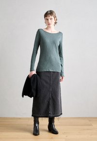 Ilgų rankovių, tamsiai žalios spalvos palaidinė iš švelnios, tekstūruotos medžiagos, derinama su juoda džinsine midi sijonu, turinčiu nuskeltas briaunas, ir juodais batais iki kelių.