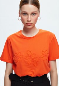 Una camiseta de algodón naranja brillante con tres diseños de flores bordados en hilo a juego, mangas cortas y un escote redondo.