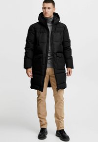 Sort polstret parka med aftagelig hætte, lynlås i lukning og frontlommer; båret over en grå turtle neck og beige bukser. Sorte sko.
