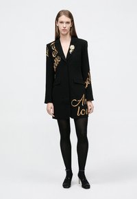 Svart blazerklänning med gulddetaljer, strukturerad design och två fickor. Har en blommig brosch på vänster sida.