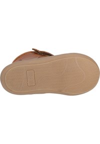 Chaussure en cuir marron avec une semelle en caoutchouc texturé couleur beige, présentant une étiquette de taille imprimée et des rainures pour une meilleure adhérence. Fermeture avec une sangle marron.