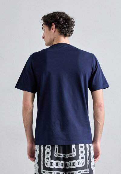 T-shirt en coton bleu marine à manches courtes et col rond, vu de dos. Un short à motifs présente de grandes graphismes de chaînes blanches.