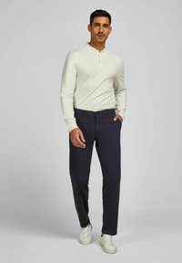 Man in een off-white langzamige henley-shirt, donkerblauwe broek en witte sneakers, staande met één hand in zijn zak.