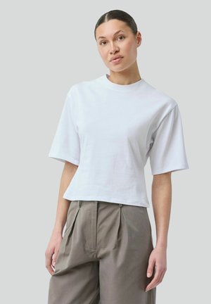 Kvinde med håret i en knold iført en enkel hvid T-shirt med korte ærmer og rund hals samt højtaljede beige plisserede bukser mod en grå baggrund.