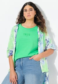 Camicia verde con la scritta "TRUE" sotto una maglietta tie-dye azzurro e verde. Jeans a vita alta blu. Capelli ricci, orecchini a cerchio d'argento.
