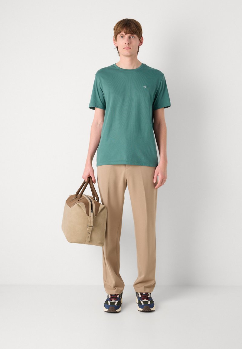 Grünes Kurzarm-T-Shirt, beige Hose und eine braune Ledertasche mit braunen Akzenten. Multicolor-Sneaker vervollständigen das Outfit.