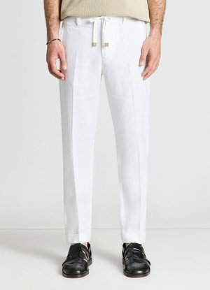 Uomo con pantaloni bianchi slim fit con coulisse e sandali in pelle neri, in piedi davanti a uno sfondo chiaro e uniforme.
