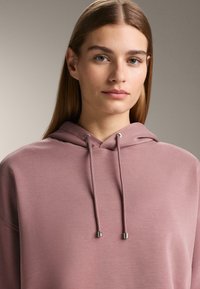 Hoodie van zachte, mauve stof; voorzien van dunne trekkoorden met metalen uiteinden en een ontspannen pasvorm. Soepele textuur, minimale details.