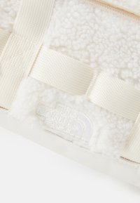 Hvit plush-bag med en myk fleeceoverflate, beige stropper og en subtil blindembossed logo. Designet er kompakt og rektangulært.