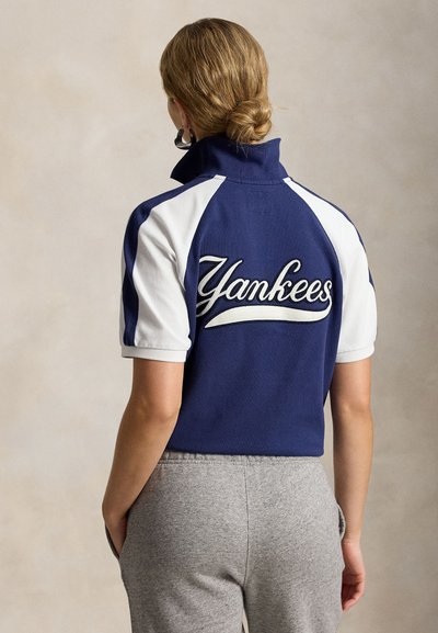 Isik juustega kuklas, kandes tumesinise ja valge lühikeste varrukatega särke, millel on taga kiri „Yankees“, kombineeritud hallide dressipükstega.