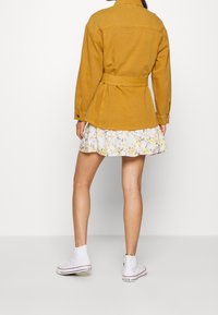 Hořčičná džínová bunda s páskem v pase, v kombinaci s bílou květinovou sukní s volánky a bílými vysokými teniskami.