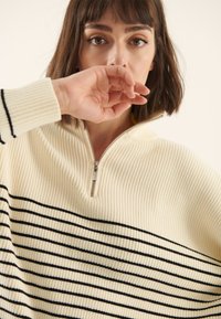 Krēmkrāsas džemperis ar melnām horizontālām svītrām, ribota tekstūra un puszipa apkakles dizains. Modelis sedz muti ar roku.