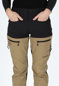 Zwarte en tan outdoor broek met ritssluitingen in de zakken, elastische tailleband en taps toelopende benen, gemaakt van duurzaam, waterbestendig materiaal.
