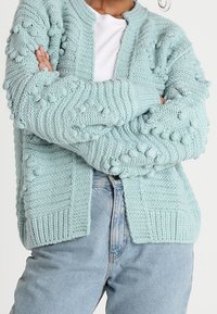 En person som bär en ljusblå bollstickad cardigan över en vit skjorta, med armarna i kors, klädd avslappnat i jeanstyg.