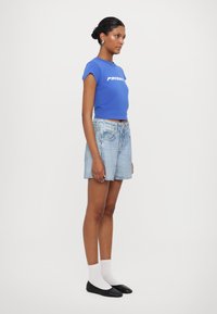 T-shirt blu corto con la scritta "PRIORITY", abbinato a pantaloni shorts in denim chiaro e ballerine nere, accessoriate con orecchini in argento.