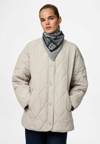 Beige quiltet jakke med knaplukning, der har oversize ærmer og sidelommer. En grå mønstret tørklæde tilføjer kontrast omkring halsen.