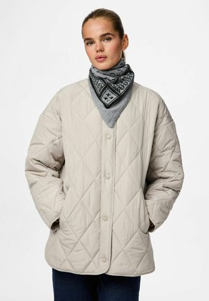 Beige quiltet jakke med knaplukning, der har oversize ærmer og sidelommer. En grå mønstret tørklæde tilføjer kontrast omkring halsen.