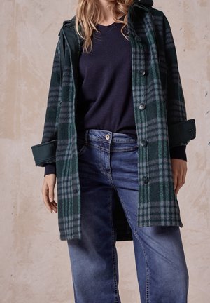 Femme portant un manteau à carreaux vert foncé et gris sur un pull noir et un jean bleu, debout contre un mur beige texturé.