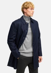 Navy sininen kevyt takki, jossa on napitettava etuosa, klassinen kaulus ja pitkät hihat, päällä harmaa poolopaita ja tummat housut.