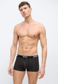 Άνδρας χωρίς πουκάμισο φορά μαύρα ανδρικά εσώρουχα με σχέδιο και ζώνη μέσης με την επιγραφή «BOSS», στέκεται μπροστά σε απλό φωτεινό φόντο.