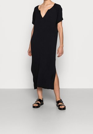 Kobieta ubrana w czarną sukienkę midi z prążkowanego materiału, z dekoltem w serek, krótkimi rękawami, rozcięciem z boku oraz czarne, odkryte sandały na platformie, stoi przy białej ścianie.