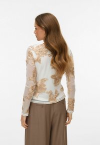 Top s dlouhými rukávy z bílé látky s béžovou květinovou výšivkou. Má průhledné rukávy a přiléhavý střih, doplněný o hnědé kalhoty s širokými nohavicemi.
