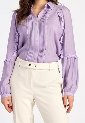 Kobieta w lawendowej bluzce z koronkowymi detalami i plisami, włożonej do wysokich, kremowych spodni z szerokim paskiem.