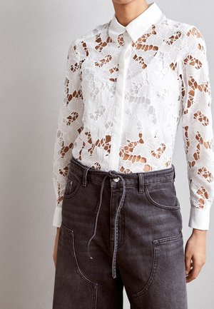 Blouse blanche en dentelle à manches longues avec motif floral et col uni, associée à un pantalon en denim noir taille haute avec une ceinture à cordon.
