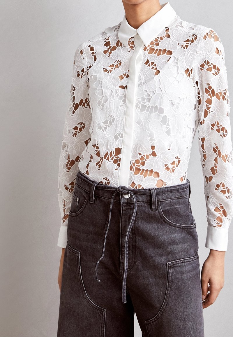 Blouse blanche en dentelle à manches longues avec motif floral et col uni, associée à un pantalon en denim noir taille haute avec une ceinture à cordon.