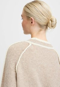 Beige gebreide trui met crèmekleurige accentstiksels, met een ronde kraag en raglanmouwen; zachte textuur zichtbaar in de stof.