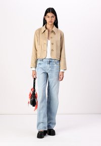 Veste cropped en suède beige avec deux poches avant, jeans taille haute à jambe large bleu clair et chaussures compensées noires. L'accessoire comprend un petit sac noir avec une écharpe rouge.