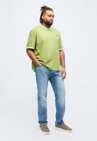 Άνδρας με ντραντλοκς που φοράει ανοιχτό πράσινο μπλουζάκι, μπλε τζιν, καφέ αθλητικά παπούτσια, στέκεται με το ένα χέρι στην τσέπη μπροστά σε απλό λευκό φόντο.