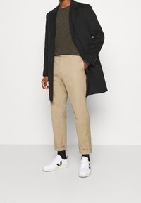 Musta pitkä takki, oliivinvihreä neulepaita, beige kapenevat housut ja valkoiset lenkkarit, joissa mustia yksityiskohtia; sileät tekstuurit ja puhtaat linjat.